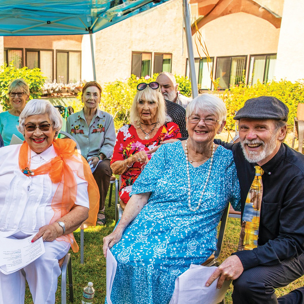A group of smiling older adults sits outdoors under a blue canopy, attending a gathering. Some hold papers, and everyone appears cheerful and dressed in colorful clothing. Greenery and buildings are visible in the background.