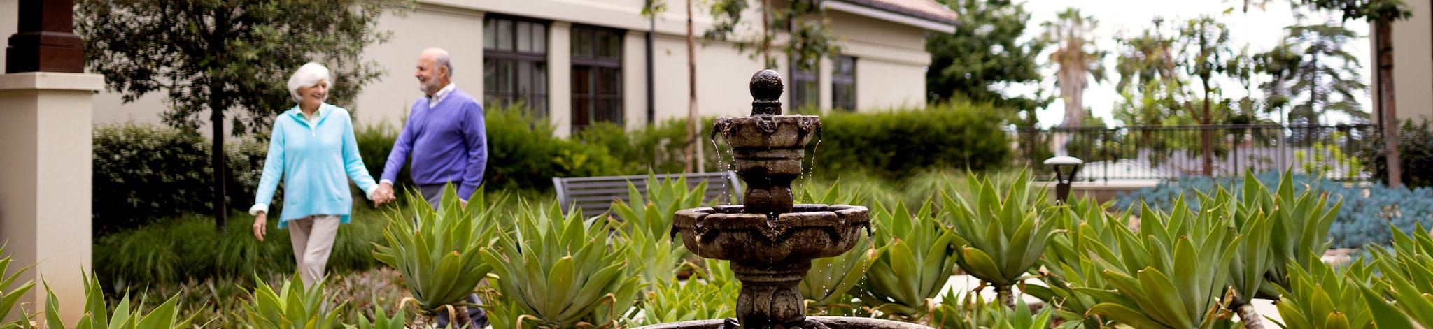 An elderly couple holds hands while walking in a lush garden near a building, with a stone water fountain in the foreground surrounded by green plants.