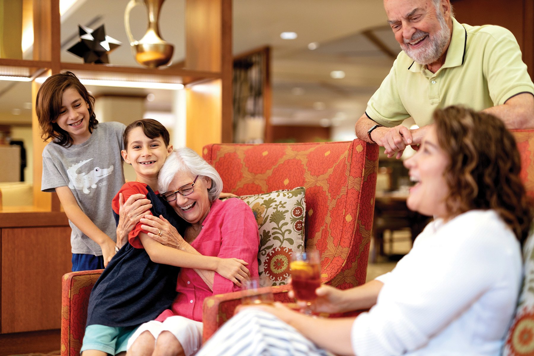 Two children hug an elderly woman sitting in an armchair, while another woman and an older man smile nearby in a warmly lit living room. Everyone appears happy and engaged with each other.