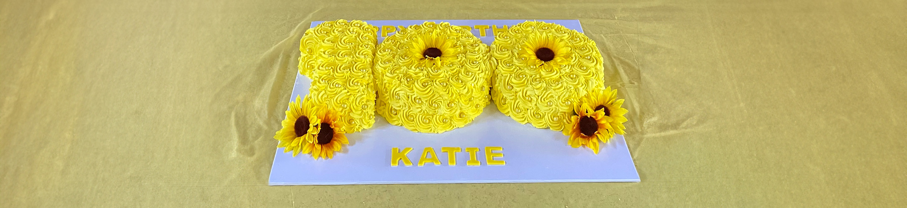 A yellow cake shaped as the number 100 with floral icing and sunflower decorations sits on a board that reads KATIE in yellow letters.