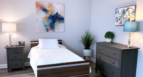 A neatly made single bed with a white textured cover is between two dark wood nightstands, each with a lamp. There are colorful abstract paintings on the light gray walls and a potted plant near a dark dresser.