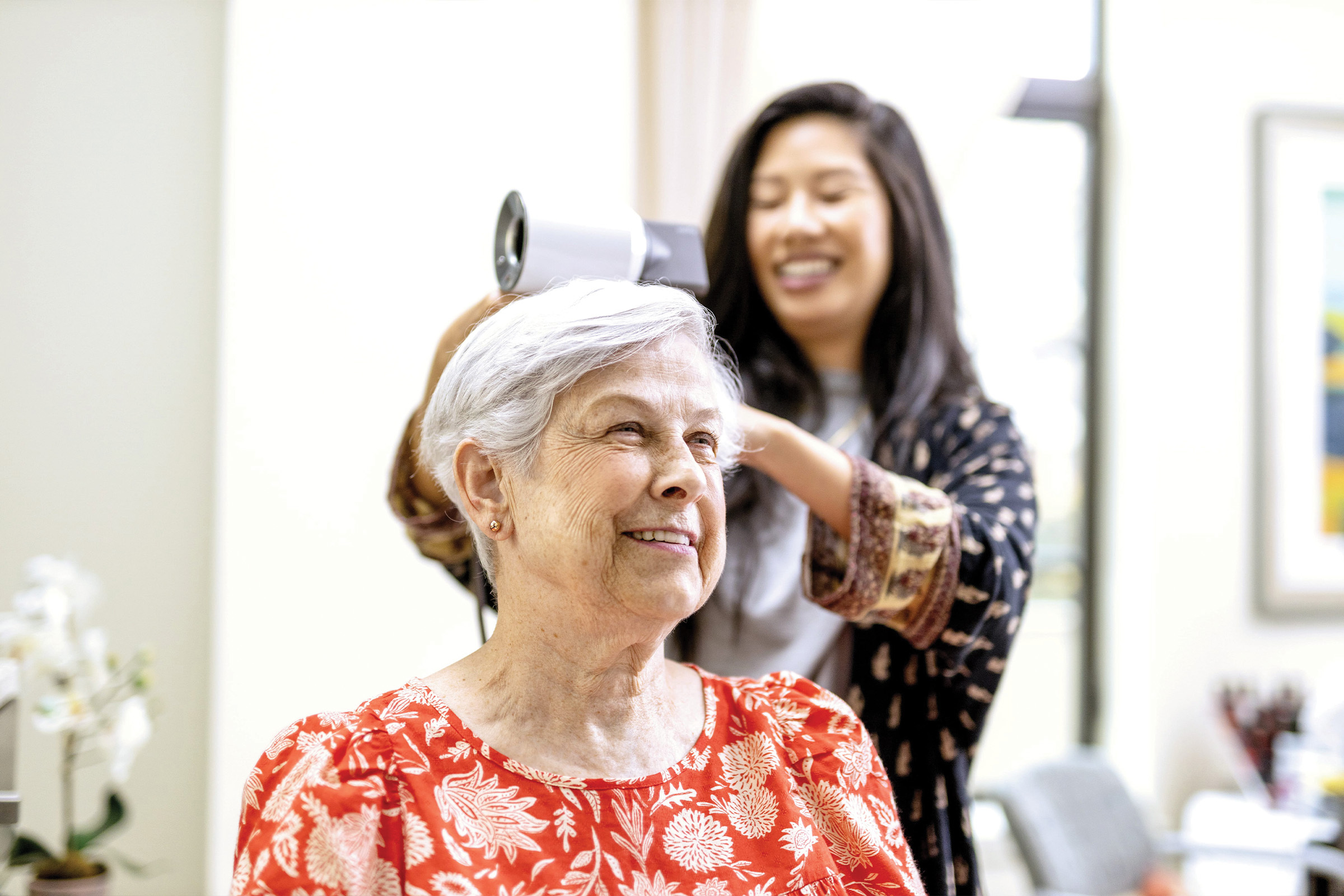 ECS-R001_Site-Update-2024_MC_Life-Plan-Communities_CL&PoM-image A smiling elderly woman with short gray hair sits as a younger woman dries her hair with a hairdryer in a bright, cheerful room.