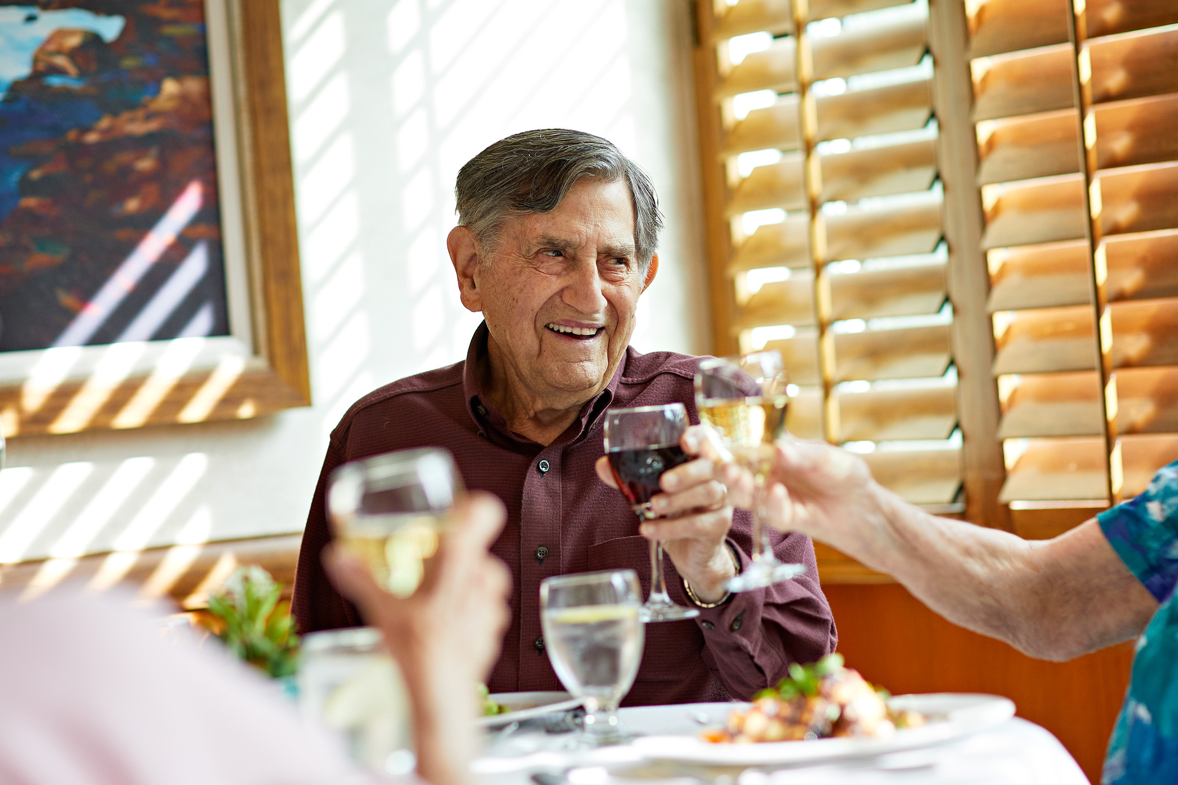 An older man smiles and raises a wine glass in a toast with friends at a sunlit restaurant table, with food and drinks in front of them and wooden blinds in the background.
