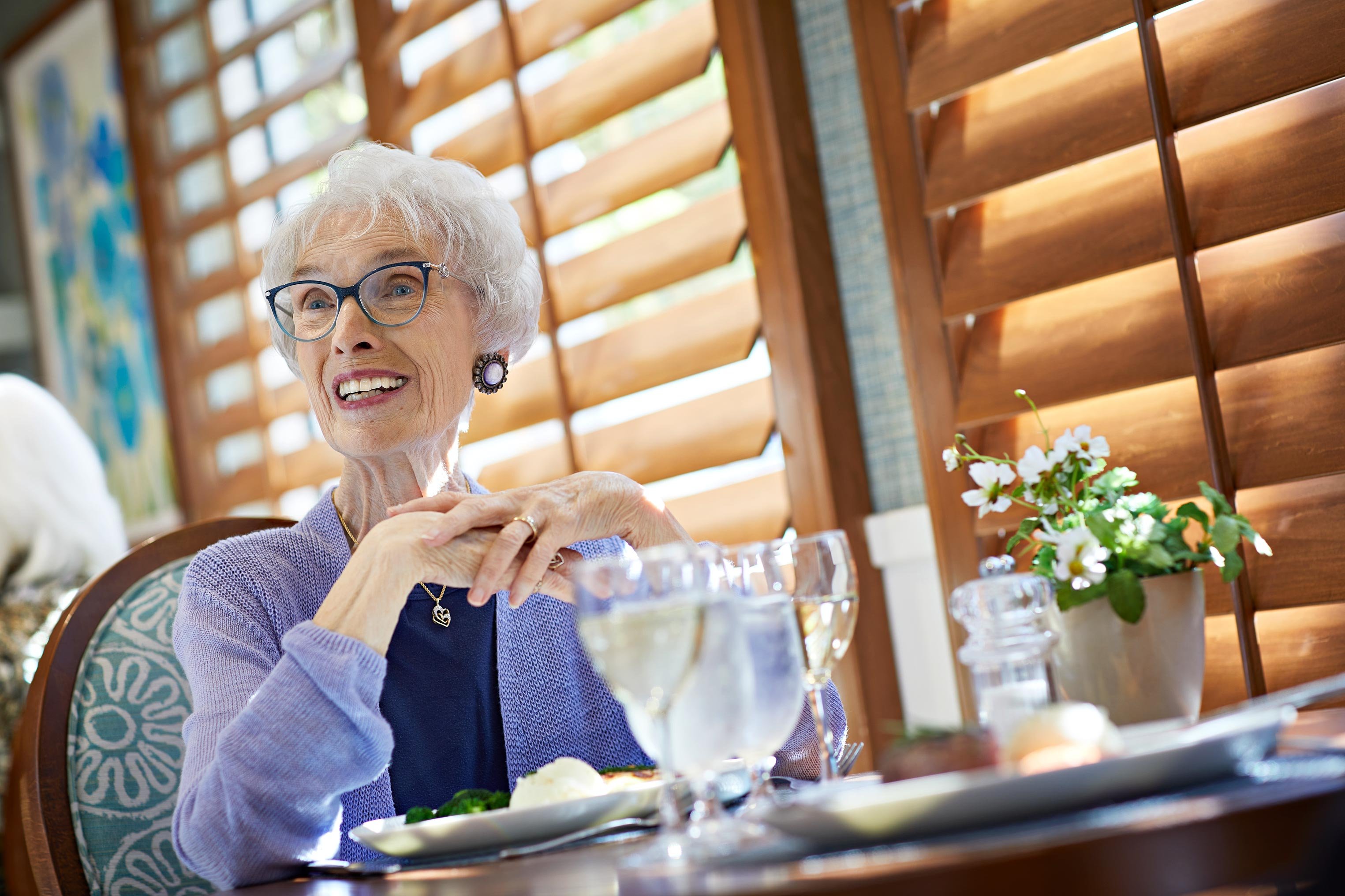 An elderly woman with white hair and glasses smiles while sitting at a dining table. She is wearing a lavender sweater and has her hands folded. Plates of food and glasses of water are on the table, with sunlight streaming through wooden blinds.