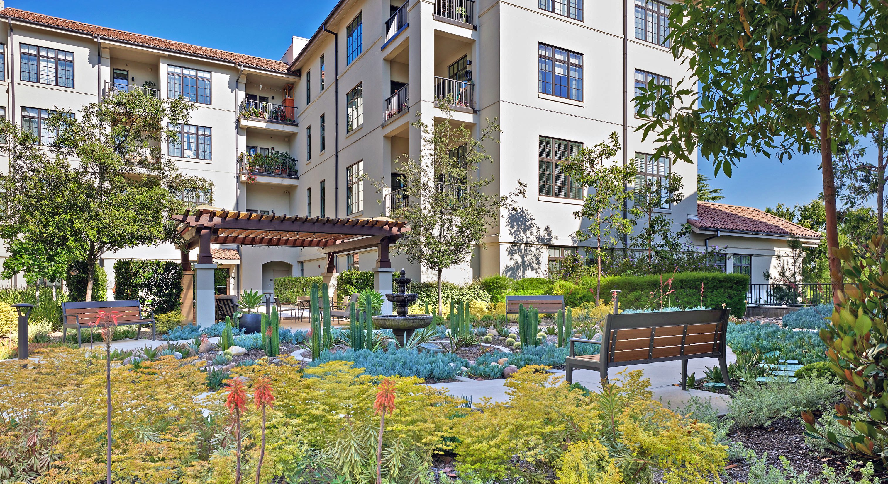 A landscaped courtyard with benches, a pergola, a water fountain, and various plants is surrounded by a multi-story beige apartment building on a sunny day.