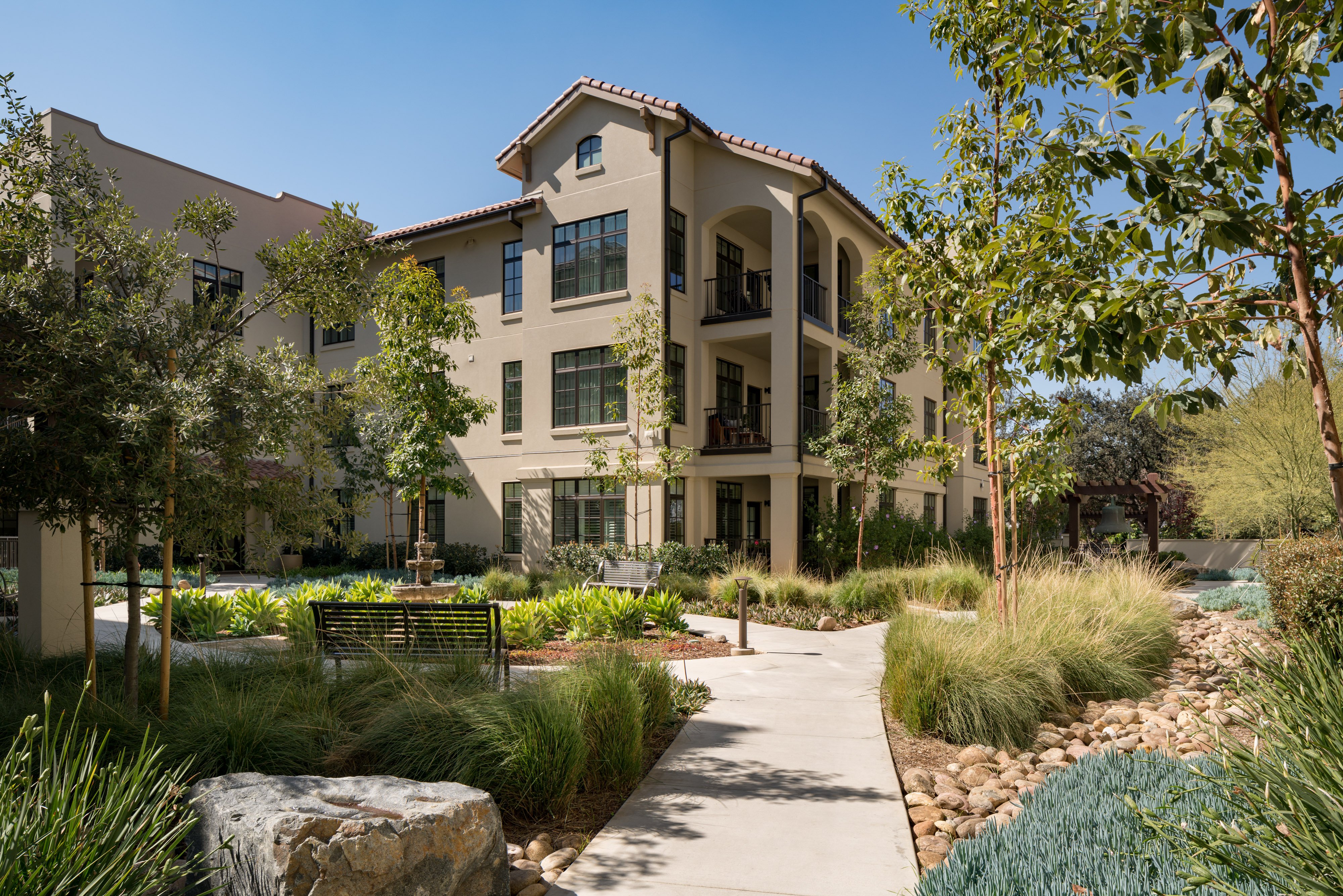 A modern, three-story apartment building with beige stucco walls, arched balconies, and a red tile roof, surrounded by landscaped gardens, trees, and a winding concrete pathway with benches.