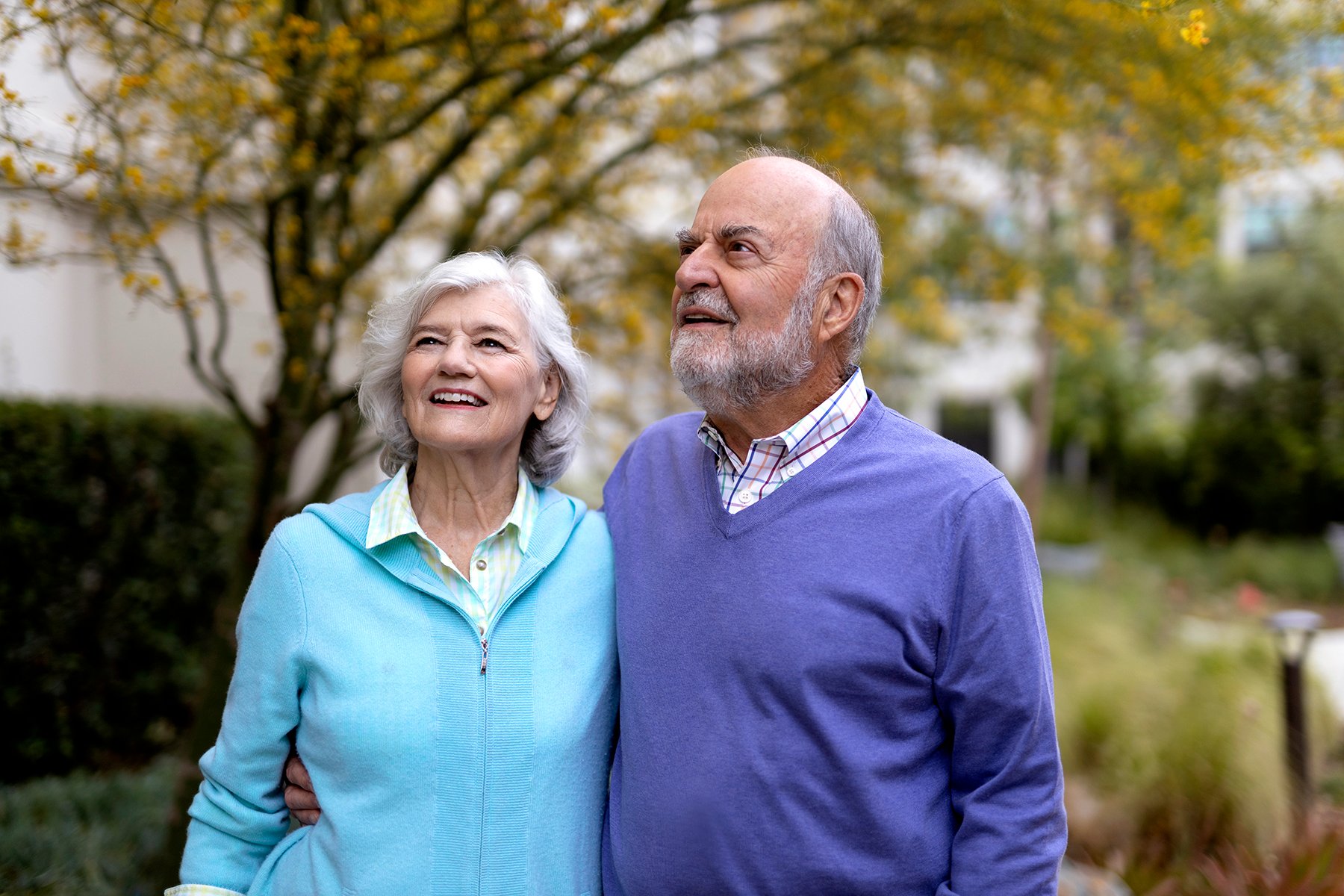An older couple stands outdoors, smiling and looking up. The woman has white hair and wears a light blue jacket, while the man has a beard and wears a purple sweater. Trees and greenery are visible in the background.