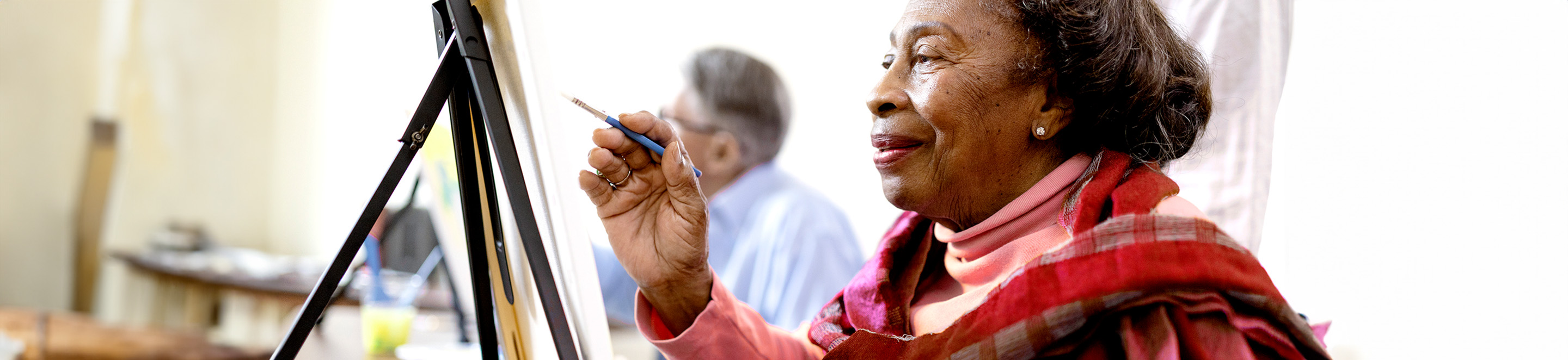 An elderly woman wearing a red plaid scarf smiles while painting at an easel. A man sits and paints in the background, both appearing to enjoy an art class in a bright room.