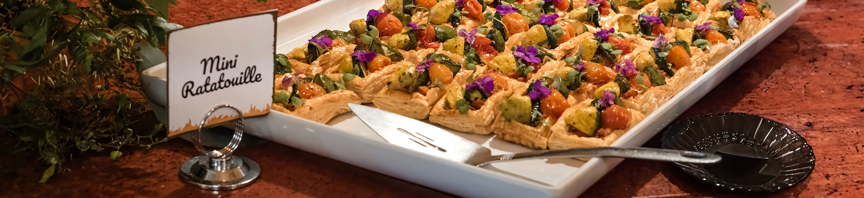 A white tray displays small servings of mini ratatouille topped with purple edible flowers. A serving spatula and a black plate are nearby. A sign reads Mini Ratatouille. The tray sits on a reddish surface.