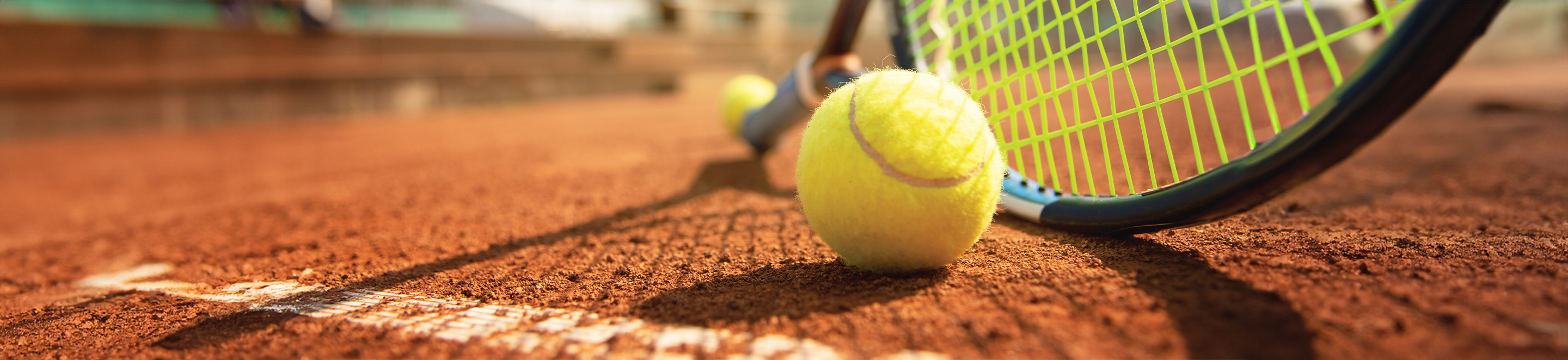 Close-up of a tennis ball resting on a clay court near the white baseline, with a tennis racket lying beside it. The scene is bright and the background is blurred.