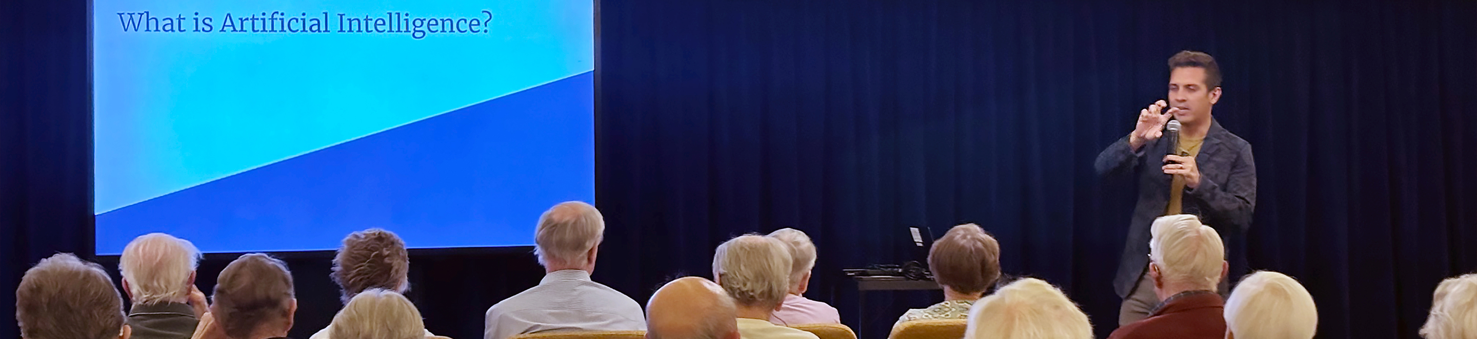 A man is giving a presentation to an audience of older adults. A large screen beside him displays the text What is Artificial Intelligence? on a blue background.