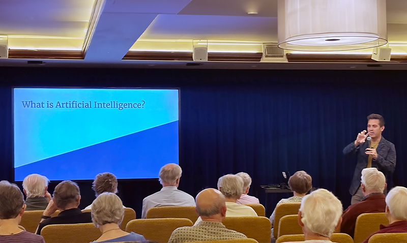 A speaker stands in front of an audience of older adults, presenting a slide titled What is Artificial Intelligence? in a conference room with a blue curtain backdrop.