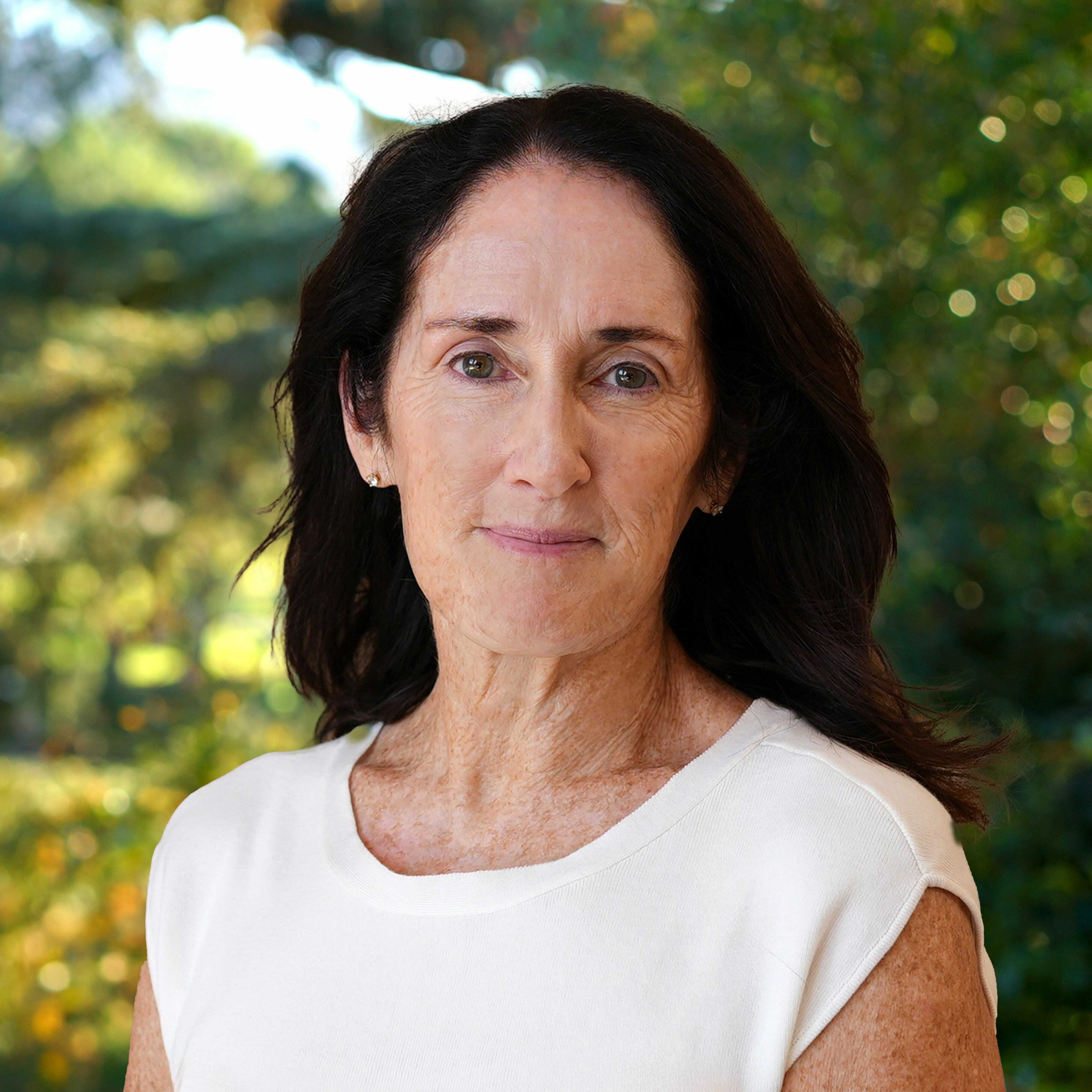 A middle-aged woman with long dark hair and a white sleeveless top stands outdoors, looking calmly at the camera with greenery and sunlight in the blurred background.