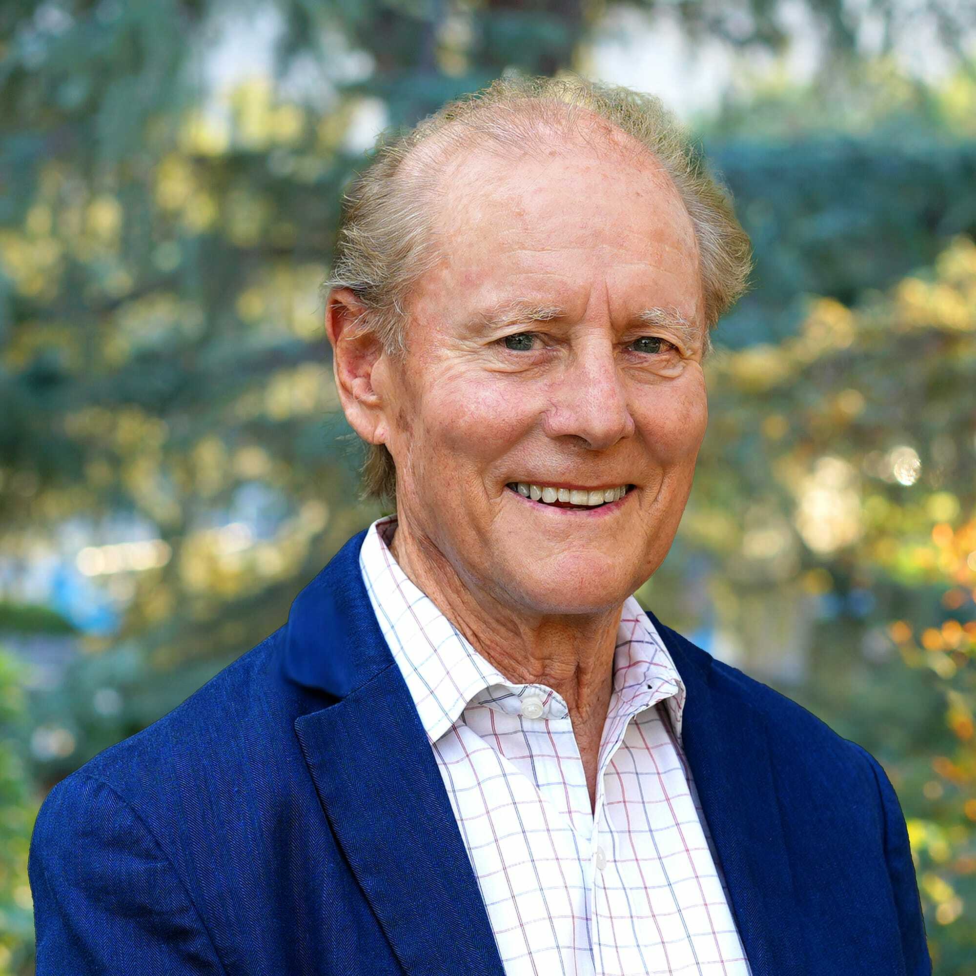 An older man with short light hair, wearing a blue blazer over a white checkered shirt, smiles outdoors with green trees and sunlight blurred in the background.