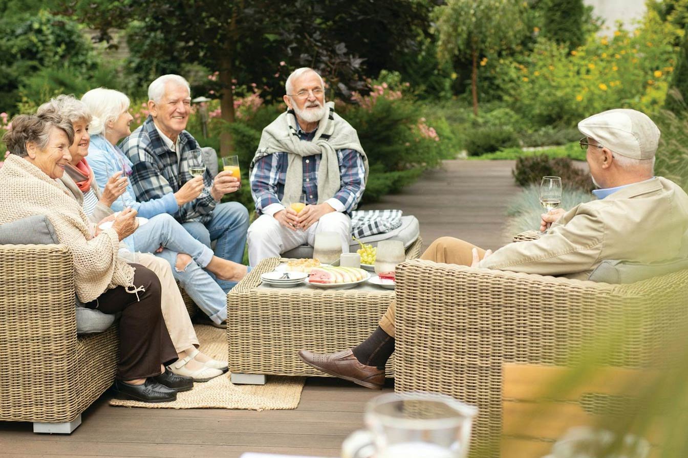 A group of six older adults sit on wicker chairs around an outdoor table, smiling and holding drinks, enjoying a relaxed conversation in a lush garden setting.