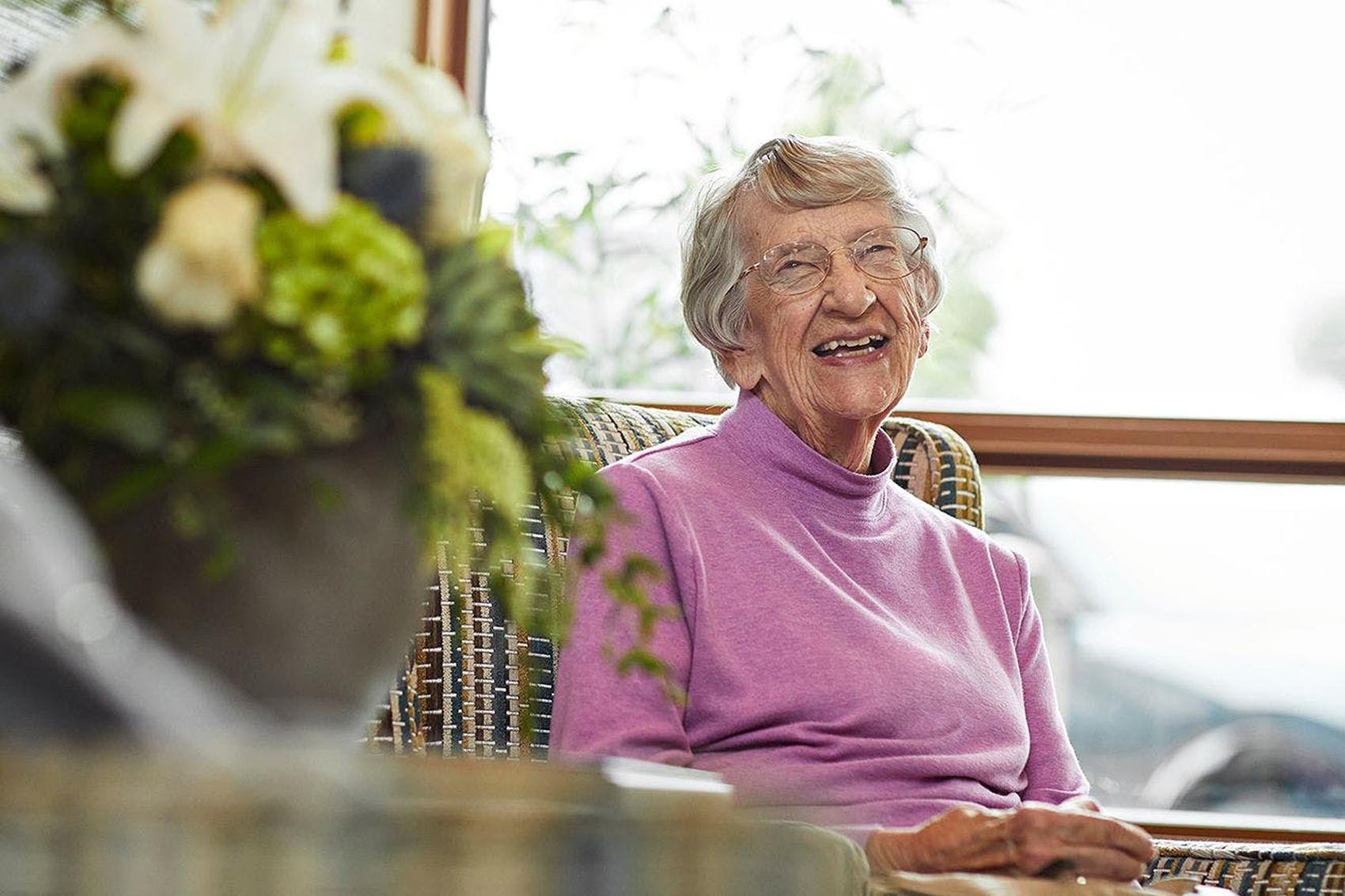 An elderly woman with short gray hair and glasses smiles while sitting in a wicker chair. She wears a light purple turtleneck. A large vase of flowers sits in the foreground near a window.