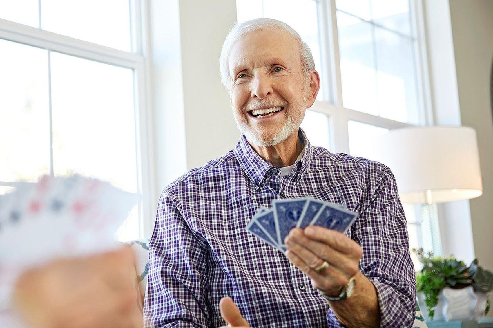An older man with white hair and a beard smiles while holding playing cards, sitting indoors by large windows. Another person’s hand with cards is visible in the foreground.