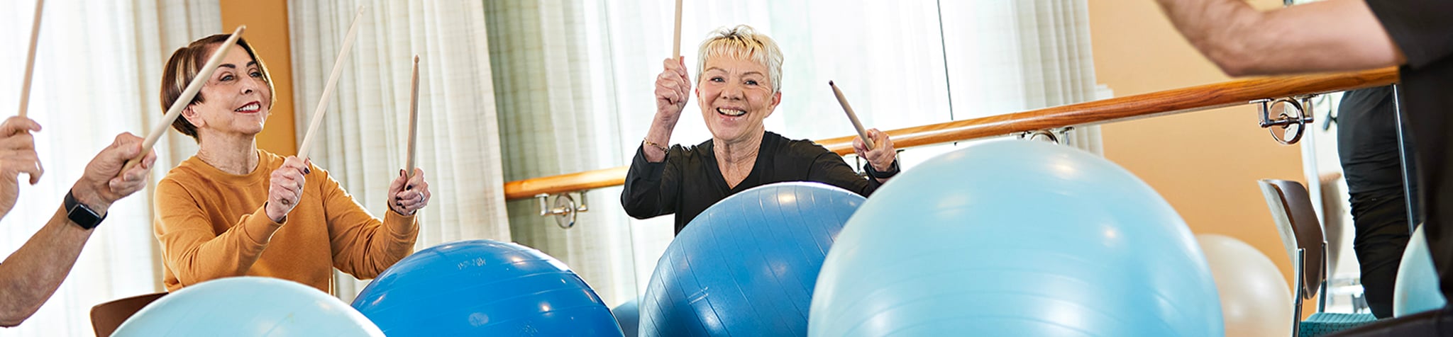 Three smiling older adults exercise indoors, using drumsticks to hit large blue exercise balls in a group fitness class.