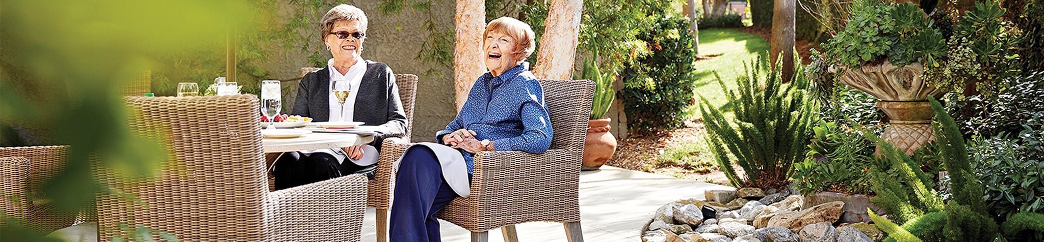 Two older women sit at an outdoor table in a garden, smiling and enjoying each other’s company on a sunny day. The setting includes wicker chairs, greenery, and a plate of food and drinks on the table.