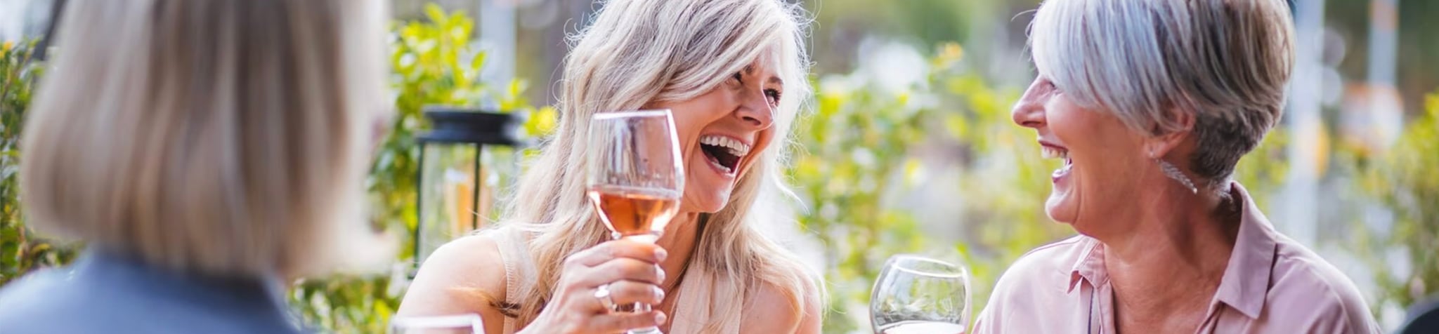 Three women with light hair are sitting outdoors, smiling and laughing together while holding glasses of wine, enjoying a cheerful moment in a sunny garden setting.
