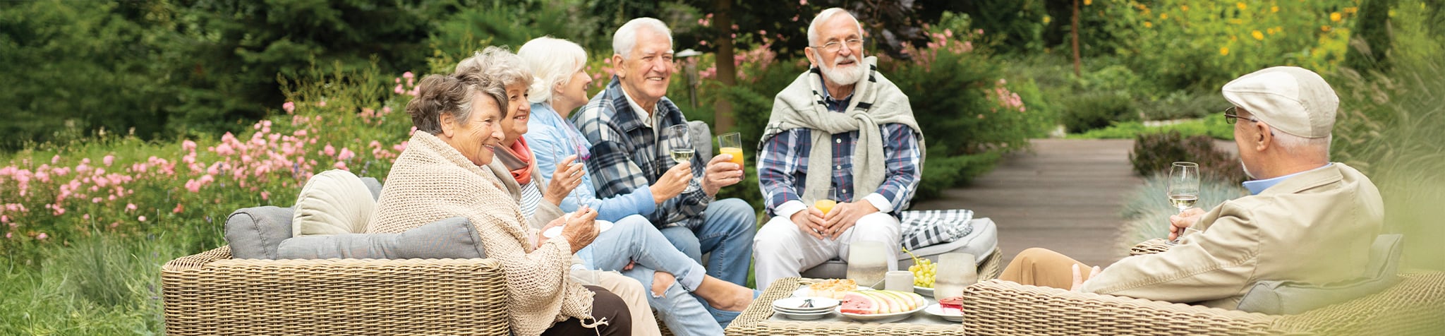A group of six older adults sit on wicker outdoor sofas in a garden, enjoying drinks, snacks, and conversation. They are smiling and appear relaxed, surrounded by greenery and pink flowers.