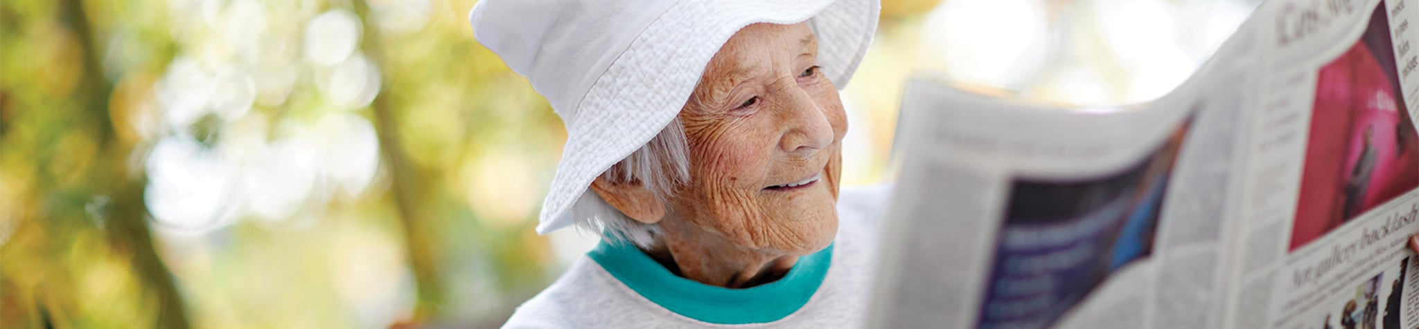 An elderly person wearing a white sunhat and light clothing smiles while reading a newspaper outdoors, with a blurred background of greenery and sunlight.