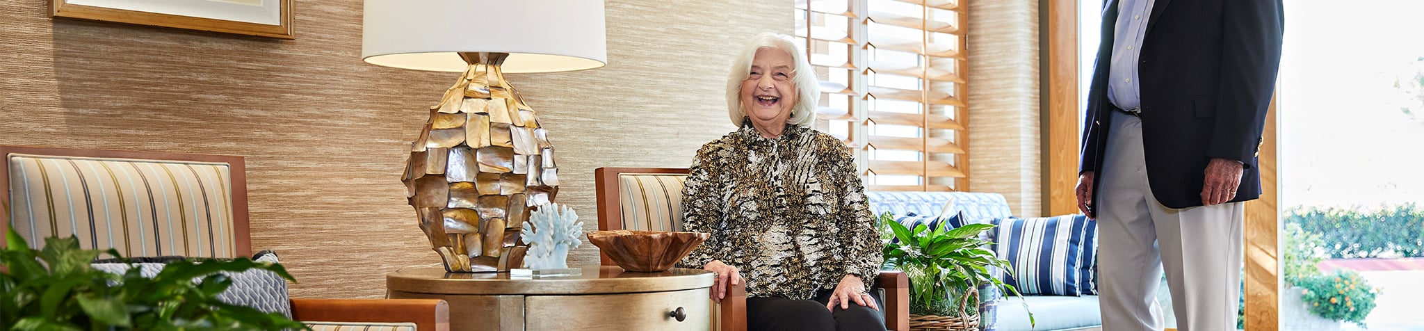 An older woman sits and smiles in a well-lit living room with modern decor, plants, and a large lamp, while an older man stands nearby, partially out of frame.