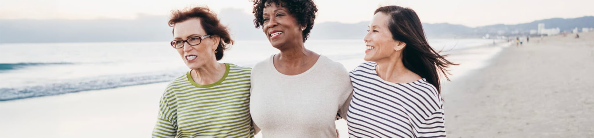 ECS-R001_Website-Hero-Updates_Dec2024_CO_Local-Area Three women walk arm in arm along a beach, smiling and enjoying each other’s company. The ocean and distant mountains are visible in the background under a clear sky.