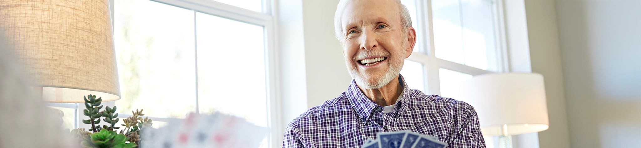 An older man with gray hair and a beard, wearing a plaid shirt, smiles while holding playing cards. He is sitting indoors near large windows with natural light and houseplants visible in the background.
