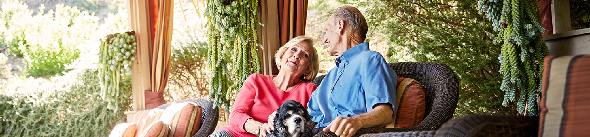 An older couple sits together on an outdoor wicker sofa, smiling and relaxing with a black and white dog on their laps, surrounded by lush greenery and hanging plants.