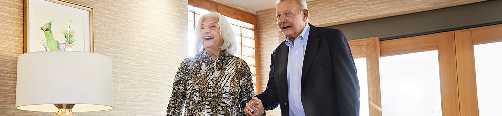 An elderly couple smiling and holding hands as they walk into a brightly lit room with wooden doors. A lamp and framed bird artwork are visible in the background.