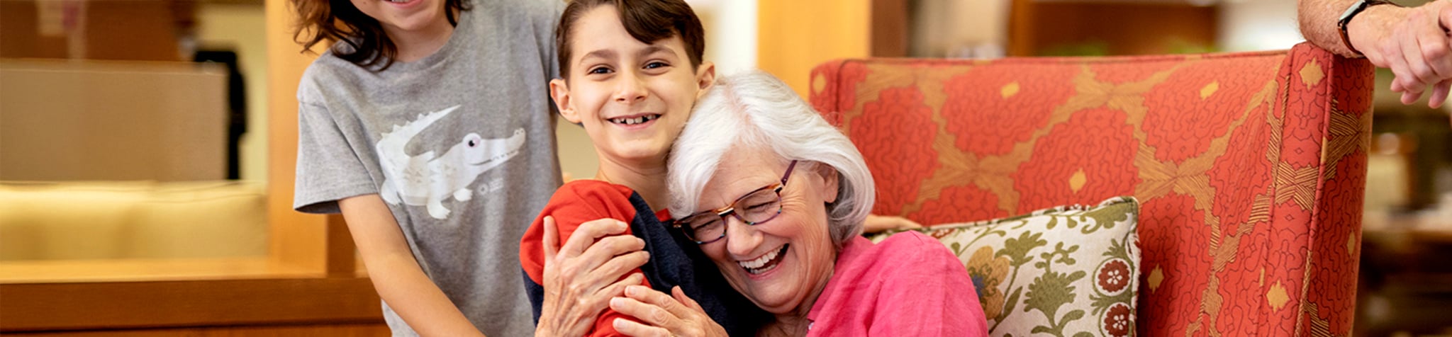 An elderly woman with white hair and glasses sits in an armchair, joyfully hugging a smiling child. Another smiling child stands behind them, all sharing a happy moment indoors.