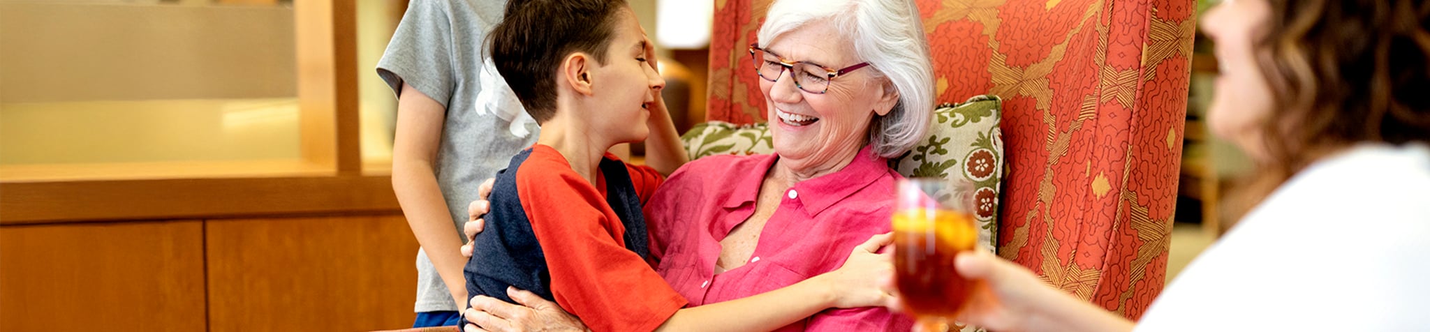 An elderly woman with white hair and glasses smiles while hugging a young child. They are seated on an armchair, with another adult and child partially visible nearby. The setting appears warm and inviting.