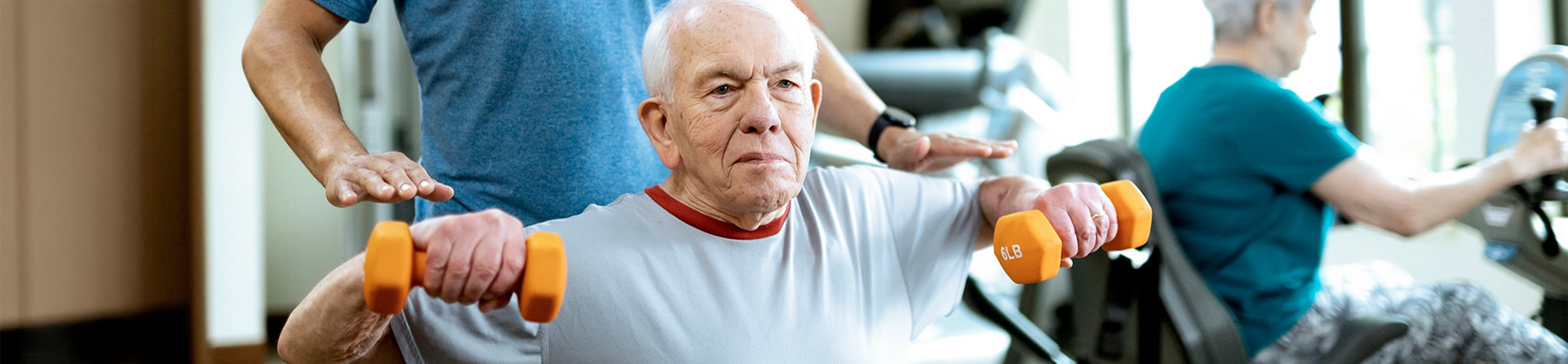 An older man lifts orange dumbbells while seated, assisted by a trainer. Another older person exercises on a stationary bike in the background. The scene is set in a bright fitness center.