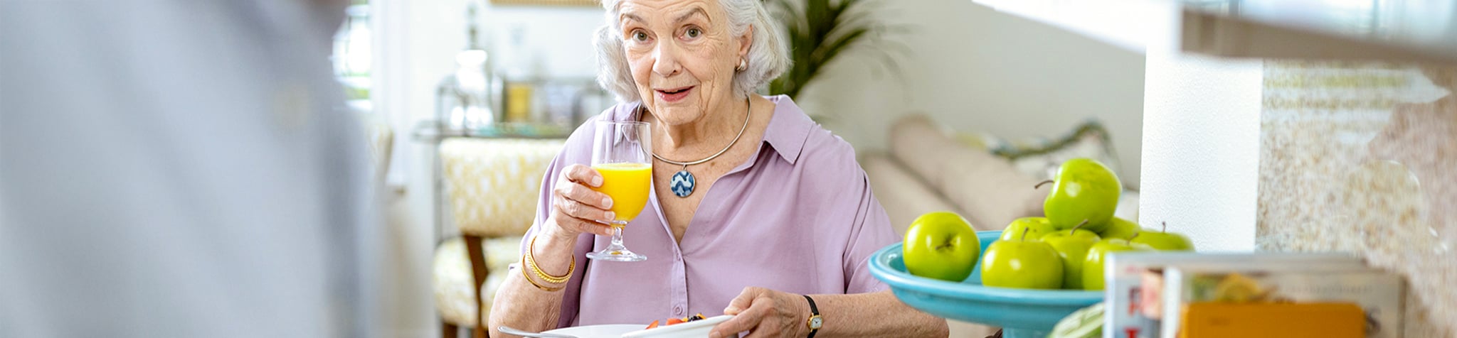 ECS-R001_Website-Hero-Updates_Dec2024_ECS_LifePlanCommunity An elderly woman with white hair, wearing a lavender blouse and a blue pendant, sits at a table holding a glass of orange juice. A bowl of green apples and magazines are visible nearby. She looks pleasantly surprised.