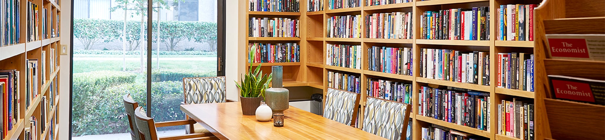 A cozy library room with bookshelves filled with colorful books, a wooden table with four chairs, potted plants, and large windows offering a view of greenery outside.