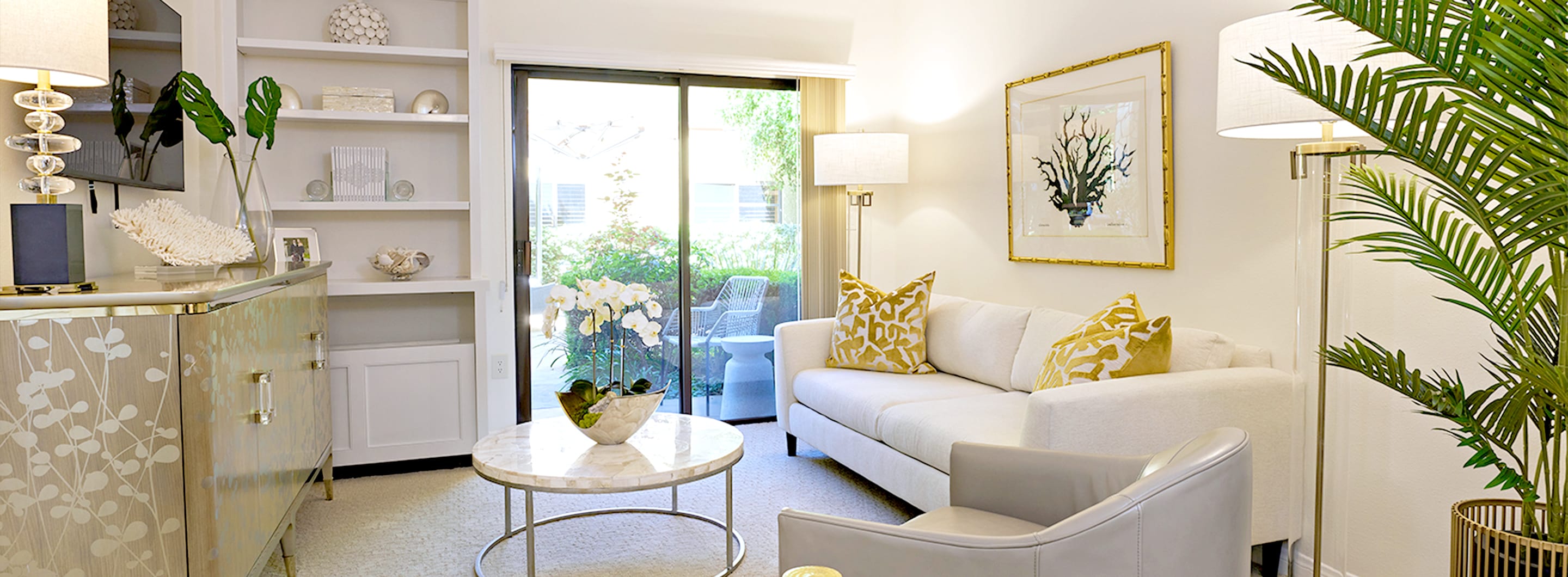 Bright living room with a white sofa, yellow patterned pillows, a gray armchair, marble coffee table, built-in shelves, large potted plant, and sliding glass doors leading to a patio with greenery outside.
