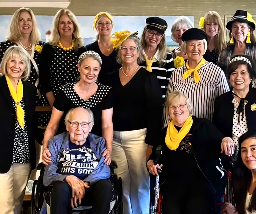 A group of smiling women, mostly dressed in black, white, and yellow with polka dot patterns, pose together. Two older individuals are seated in front, one in a wheelchair, both also smiling.