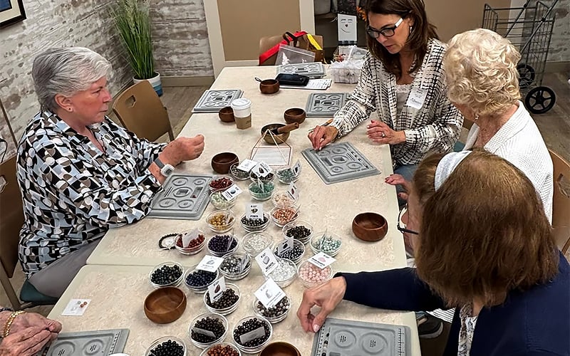 Four women sit around a table making jewelry with assorted beads in small bowls. Mats, tools, and supplies are spread out, and the group appears focused and engaged in their craft activity.