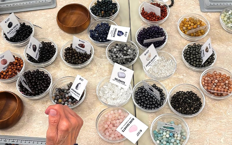 A hand reaches toward small bowls filled with various colorful gemstone beads, each labeled with a card identifying the stone type, all arranged on a beige tabletop.
