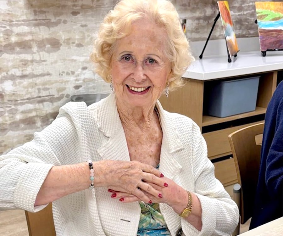 An elderly woman with curly blond hair and a white textured jacket smiles while sitting at a table, her hand resting on her chest. Paintings and art supplies are visible in the background.