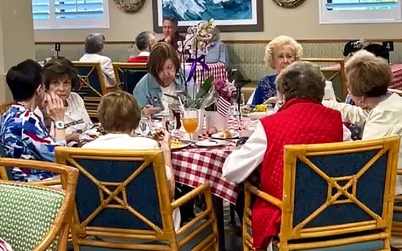 A group of elderly women sit around a table with a red-and-white checkered tablecloth, enjoying a meal together in a brightly lit dining area. Other diners are visible in the background.