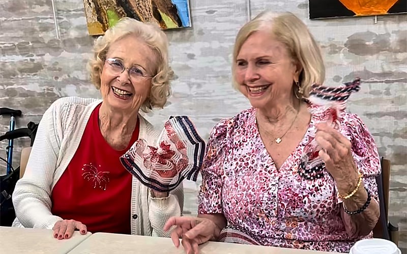 Two smiling elderly women sit at a table holding red, white, and blue patriotic decorations. One wears a red shirt, the other a pink patterned dress. Artworks hang on the wall behind them.