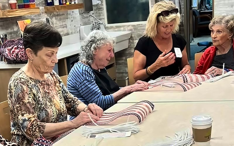 Four older women sit at a table, weaving or crafting together with striped fabric. One woman holds a tool, while others focus on their work. A coffee cup and supplies are on the table in a cozy, well-lit room.