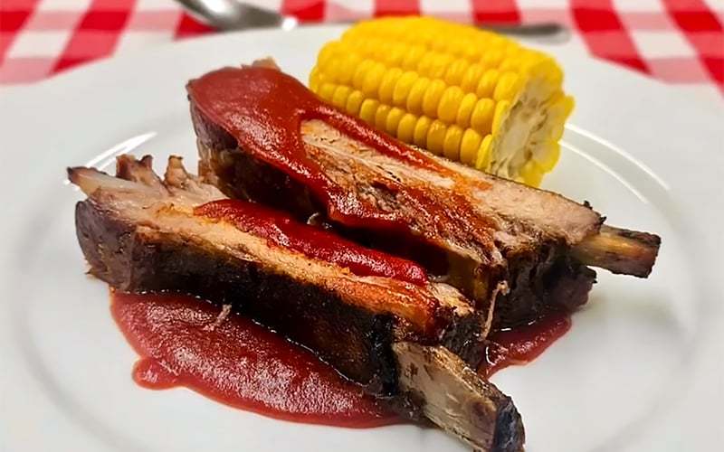 Two barbecue pork ribs topped with barbecue sauce are served on a white plate alongside a piece of corn on the cob, with a red and white checkered tablecloth in the background.