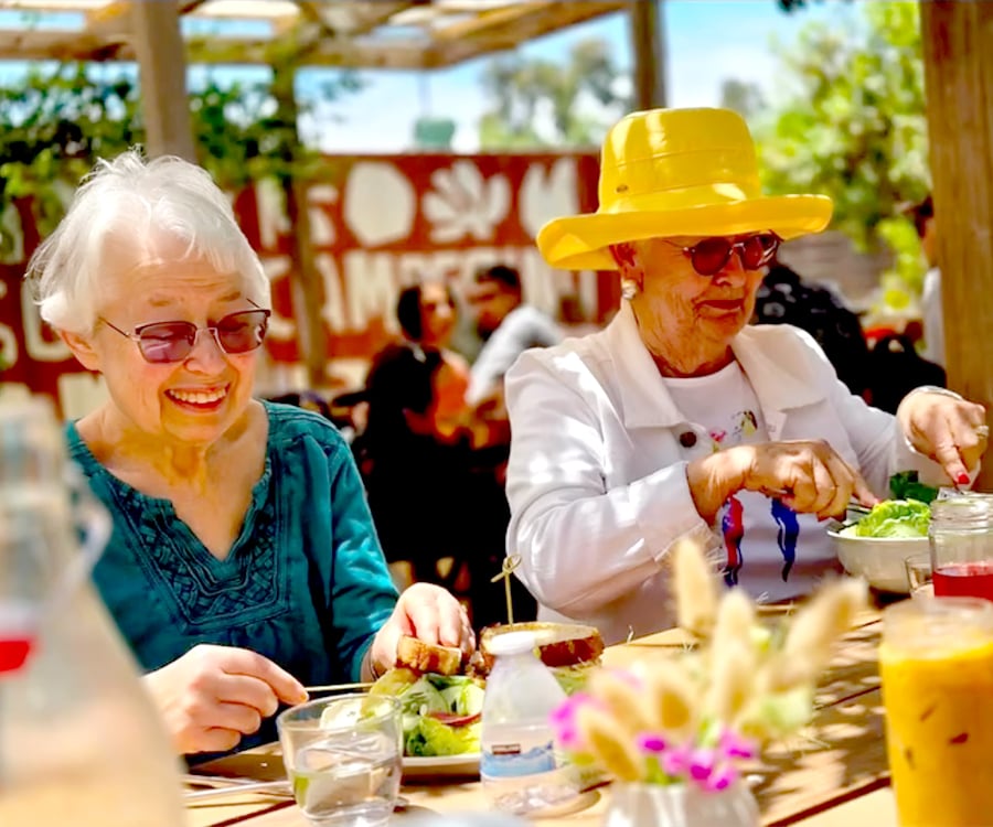 Two elderly women sit at a sunlit outdoor table, smiling and enjoying a meal together. One wears a turquoise top and glasses, the other a bright yellow hat and white jacket. There are salads, drinks, and flowers on the table.