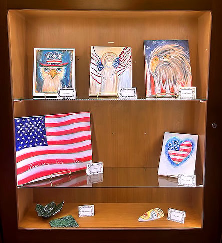 A display case with patriotic art, including two eagle paintings, an angel with American flag wings, a painted American flag, a heart with a flag pattern, and small ceramic pieces on two shelves.
