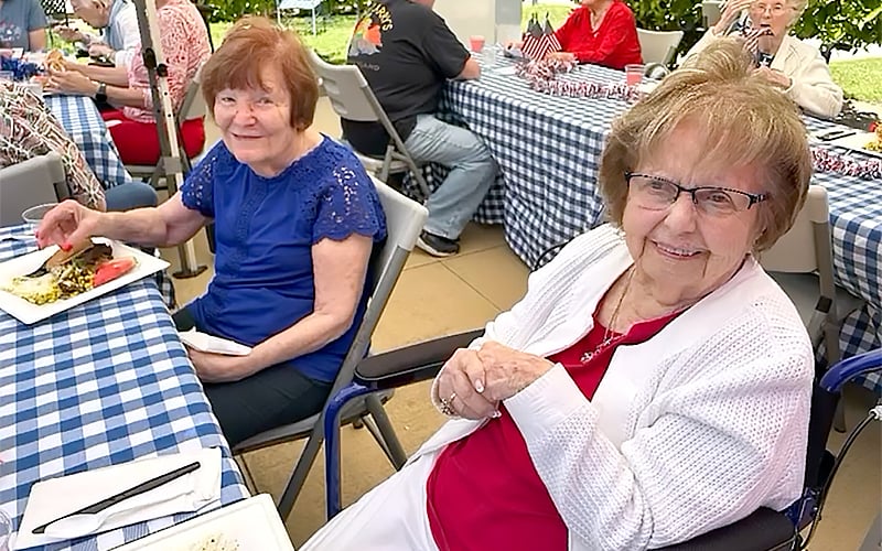 Two elderly women sit at a table with a blue-and-white checkered tablecloth, smiling at the camera. Other people are eating and talking at similar tables in the background, outdoors on a sunny day.
