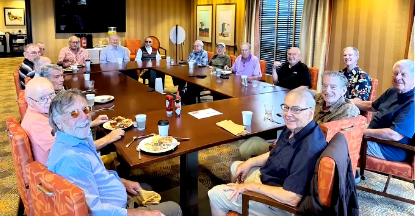 A group of older adults sit around a large square table in a warmly lit room, smiling at the camera. Plates of food and drinks are on the table, suggesting a social gathering or meal.