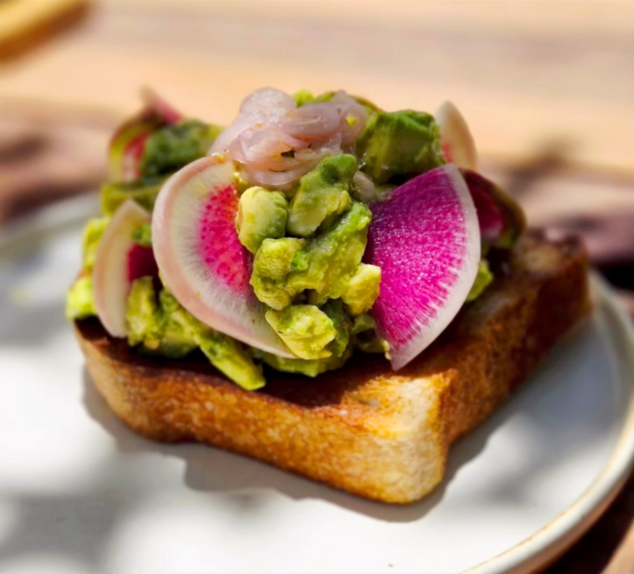 A slice of thick toasted bread topped with chunky avocado, thinly sliced vibrant watermelon radishes, and pickled onions, served on a white plate with a blurred outdoor background.